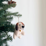 A charming felt beagle Christmas ornament with a tiny golden bell hangs gracefully from a green Christmas tree adorned with pine cones. This handcrafted felt beagle features a predominantly white body with brown and dark brown patches on its head and ears. The soft texture and jute string for hanging add a joyful touch to holiday decor.