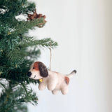 A charming felt beagle ornament, needle felted and adorned with a gold bell, hangs on a lush Christmas tree, nestled among green needles. This festive beagle features a cream-colored body with brown markings, a black nose, and a jute string for hanging.