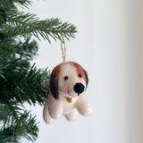 A needle felted beagle ornament, crafted from soft wool felt, hangs on a Christmas tree with green needles, set against a minimalist white backdrop. This charming beagle features a cream-colored body, brown head markings, and floppy brown ears. A golden bell is around its neck, with a jute string attached on top for hanging.