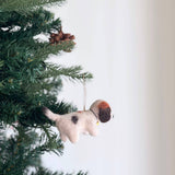 A charming felt beagle ornament, adorned with a small golden bell, hangs on a festive Christmas tree. The unique beagle ornament has a fluffy, off-white body, a distinctive black spot, and a brown patch on its head, with a delicate cotton string for hanging. Pine needles and brown pine cones surround it, evoking holiday warmth and joy.