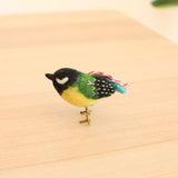 A vibrant 3D felt bird ornament, crafted from wool, rests on a light-colored wooden surface, with a green plant softly blurred in the background. This unique needle felted bird features a black head with a white cheek, a green back with stitching, a yellow body, and a black tail with white dots and a light blue tuft. Thin brown legs and a red and white hanging string complete this charming Christmas ornament.