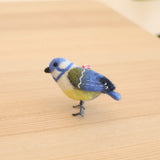 A charming handmade needle felted blue tit ornament rests on a light brown wooden surface, set against a soft, neutral backdrop. This 3D Christmas ornament vividly mimics a blue tit bird, featuring fluffy blue, white, and yellow felt with subtle stitching, a small black beak, and gray legs. A red and white string is attached for hanging.