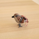 An organic wool felt sparrow ornament rests on a light wooden surface. This three-dimensional handmade felt sparrow features brown and white felt with distinctive black eyes, a black beak, and white and black chest stripes. A red and white cotton string is attached, making it a charming Christmas tree ornament.