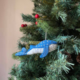 A handcrafted blue Minke whale felt ornament hangs gracefully on a festive Christmas tree, adorned with green needles, red berries, and pine cones. This needle felted ornament, made from organic wool, features a blue upper body with white dots, a white lower body with blue stripes, and blue felt fins, all detailed with white stitching. A white cotton string is attached for hanging, enhancing the joyful holiday display.