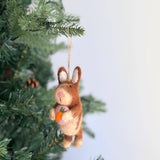 A handmade felt bunny ornament, holding a heart, hangs charmingly from a Christmas tree branch adorned with green needles and pine cones. Crafted from organic wool, the bunny is predominantly brown with lighter accents on its ears, paws, and belly, and features a pink nose. It sweetly holds a pink heart, adding a whimsical touch to the festive holiday decor.