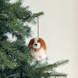 A handcrafted felt Cavalier King Charles Spaniel dog Christmas ornament hangs from a subtly lit Christmas tree branch. This brown and white felt ornament is meticulously shaped with distinctive markings and features. A delicate golden bell hangs from a black string around its neck, adding a festive touch to the warm seasonal scene, suspended by a jute string.