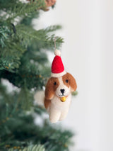 A handmade felt Cavalier King Charles Spaniel dog ornament hangs on a vibrant green Christmas tree. The brown and white dog features a fluffy tail, a golden bell around its neck, and a red Santa hat with a white pompom and band atop its head. A jute string is attached for hanging.