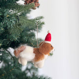A charming handmade felt Cavalier King Charles Spaniel dog ornament hangs on a Christmas tree, radiating a joyful, festive atmosphere. This brown and white felt dog wears a red Santa Claus hat with a white pompom and brim, and a gold bell around its neck. It hangs from a rustic jute string.