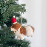 A Cavalier King Charles Spaniel dog felt ornament hangs delicately from the branches of a festive Christmas tree. This handmade felt ornament is crafted from soft felt, showcasing white and brown fur. It wears a red and white Santa hat and has a golden bell around its neck, evoking a joyful, celebratory mood.