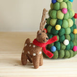 A brown wool felt reindeer Christmas ornament stands on a light-colored wooden surface, next to a decorative Christmas tree. This festive felt reindeer features white spots, a bright red scarf, and small, textured antlers, adding a whimsical touch to holiday decor.