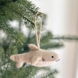 A playful needle felted shark ornament hangs from a green Christmas tree branch, set against a blurred white background. This unique Christmas tree ornament is light brown with a darker upper body and a lighter lower section. It features a dorsal fin, two side fins, a tail fin, a black eye, and black stitched details for the mouth. A brown jute string is attached, ready for display.