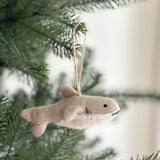 A playful felt shark Christmas ornament hangs on a lush green Christmas tree. This soft, light brown handmade felt shark ornament features darker accents defining its form, complete with a dorsal fin, side fins, and a tail. Black lines indicate gills, and a stitched white grin reveals pointy teeth. A jute string allows for hanging this charming felted ornament.