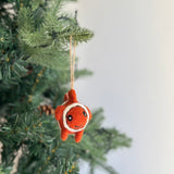 A playful wool felt clownfish ornament hangs from a green Christmas tree branch against a soft background. This charming handmade ornament features a bright orange body with white accents and a stitched smile, suspended by a jute string. Its endearing design captures the spirit of a clownfish, adding a festive touch.
