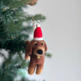 A handcrafted felt dachshund Christmas ornament hangs festively on a Christmas tree. The brown felt dachshund features lighter brown legs, a black nose, and detailed facial features. It is adorned with a red and white striped Christmas hat and a small golden bell, suspended by a jute string.