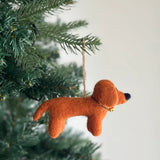 A handmade felt dachshund ornament hangs suspended from a festive Christmas tree branch. This charming brown dachshund features a distinct black nose, adding a joyful and cozy touch to holiday decorating.
