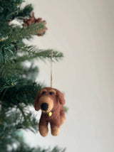A wool felt Dachshund Dogs ornament hangs on a Christmas tree. This unique Christmas ornament features a white felt frame encasing four adorable dachshunds in cream, khaki, brown, and black, alongside a playful felt bone at its center. It is suspended by a jute string, evoking a warm and festive mood.