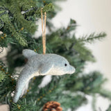 A blue-grey and white dolphin felt ornament hangs gracefully on a lush green Christmas tree. This unique Christmas tree ornament features a streamlined, curved shape with a pointed snout and a small black eye. Its dual-tone design includes a light blue-grey upper body and a creamy white underside, with a jute string for hanging.