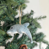 A light blue felt dolphin ornament hangs joyfully from a Christmas tree, nestled among green needles with a pine cone. This handmade felt ornament features a smooth texture, a streamlined body with a dorsal fin, a pointed snout, a rounded tail, and dark eyes, adding a whimsical touch. A jute string allows for easy hanging.