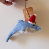 A handcrafted felt dolphin ornament is gently held by a hand against a muted background. The light blue dolphin features a white underbelly, a black eye, and a stitched mouth, crafted from soft wool felt. A festive red Santa hat with a fluffy white pompom sits jauntily on its head, and it hangs from a jute string attached to a light brown product tag.