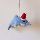 A needle felted dolphin ornament hangs by a jute string against a pale background. This light blue dolphin features a white underbelly and a charming red and white Santa hat. Its small black dot eye and soft felt texture highlight its handcrafted appeal, creating a whimsical, festive mood.