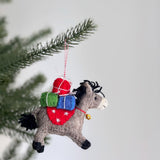 A whimsical grey felt donkey ornament carrying a stack of colorful gifts hangs from a cotton string, partially framed by a green Christmas tree branch. This delightful handmade felt ornament features the grey donkey with black hooves and mane, a red saddle blanket adorned with white snowflakes, and a golden bell with a red string around its neck. Three red, green, and blue felt gifts are tied with white strings on its back.
