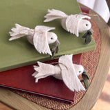 Three handcrafted wool felt dove ornaments, each with a delicate olive twig in its beak, are artfully arranged on a stack of red and green books. The pure white felted doves feature a jute string for hanging and are displayed on a round, woven surface, creating a tranquil and serene atmosphere.