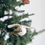 A handcrafted wool felt dove ornament with an olive twig in its beak hangs on a snow-covered Christmas tree branch, creating a serene, festive scene. The pure white dove ornament, suspended by a jute string, features a green felt olive twig. This unique Christmas ornament brings a peaceful, winter atmosphere.