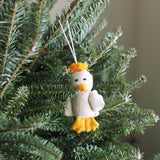 A handmade felt duck ornament is nestled among the green needles of a Christmas tree. This charming duck features a cream-colored body, black eyes, an orange beak, orange feet, and a tuft of orange hair. A white cotton string is attached for easy hanging.
