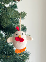 A needle felted Easter chick ornament hangs from the vibrant green branches of a Christmas tree, radiating playful holiday charm. Crafted from soft white wool felt, this cheerful chick features black eyes, an orange beak, a bright red comb, and distinctive bow-like wings. Orange felted feet peek from its bottom, with a sturdy hemp string attached for hanging.