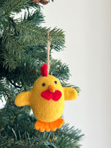 A handmade felt Easter Chick ornament hangs on a lush green Christmas tree. The whimsical yellow, egg-shaped chick has a vibrant red comb and wattle, orange feet, and small black eyes. Crafted from soft wool felt, it features hand-stitching and a sturdy hemp string for hanging, creating a cheerful holiday spirit.