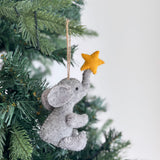 A handcrafted felt elephant ornament featuring a star hangs on a festive Christmas tree. This gray felt elephant is posed in a seated position with its trunk reaching upwards, holding a small yellow star at its tip. A jute string loop is attached for easy hanging, highlighting its unique handmade aesthetic.