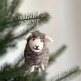 A fluffy wool felt sheep ornament hangs from a vibrant green Christmas tree branch. This charming sheep features a gray wool felt body, with a cream-colored face and inner ears. Its face has stitched black eyes and a smile, complemented by three white wool felt legs. A jute string is attached at the top for hanging.