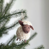 A fluffy sheep felt ornament hangs on a green Christmas tree branch, set against a bright, neutral background. This charming wool felt sheep ornament features a cream-colored, woolly body and a dark brown felted face with small, stitched eyes and mouth. A jute string is attached for easy display, creating a cozy and festive look.