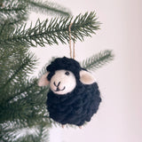 A fluffy sheep felt ornament hangs on a Christmas tree branch, creating a cheerful, festive mood. This handmade wool felt ornament features a black sheep with a white face, fluffy ears, a stitched mouth, and button eyes. Its textured wool adds a cozy appeal, enhancing the classic Christmas scene.
