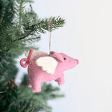 Pink felt Flying Pig ornaments hang from a green Christmas tree branch, evoking a festive atmosphere. Each whimsical felted Flying Pig ornament features white wings and a curly tail, suspended by a jute string against a plain white background.