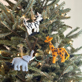 A handcrafted felt giraffe ornament hangs playfully from a Christmas tree branch by a jute string. The unique felt giraffe ornament is textured in orange with distinctive brown spots and features a small yellow tail.
