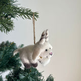 A charming wool felt goat ornament hangs from a Christmas tree branch, presented against a soft white background. This needle-felted goat features a predominantly white body with dark brown hooves and distinctive striped ears. A golden bell adorns its neck, and a jute string loop is at the top for easy hanging, evoking a cozy, festive mood.