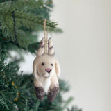 A needle-felted goat Christmas ornament made from organic wool hangs festively on a Christmas tree. This charming goat ornament features a cream body, dark brown hooves, gray ear accents, a pink nose, and a hand-stitched mouth. A jute string is attached for easy hanging.