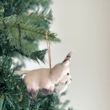 A needle-felted goat ornament hangs on a vibrant green Christmas tree. This charming wool felt goat features a creamy white body, dark brown legs and ears, and a fluffy white beard, ready to adorn any festive display with its simple jute string hanger.