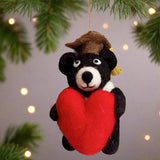 A charming graduation black bear felt ornament, clutching a vibrant red heart, hangs from a festive pine branch with soft bokeh lights in the background. This handcrafted felt ornament features a black bear adorned with a brown graduation cap and a gold tassel. The unique Christmas ornament is suspended by a delicate gold string, perfect for celebrating accomplishments.