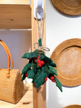 A festive felt holly sprig ornament hangs from a white hook on a wooden shelf post in a naturally lit, warm-toned interior. This handmade ornament features deep green felt leaves and bright red felt berries, bound by a jute string with a bell and a loop for hanging. A woven bag and straw hats in the background complete the natural, cozy setting.