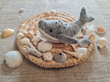 A gray wool felt Humpback Whale ornament rests on a woven straw mat, surrounded by various seashells and a burlap background. This handcrafted felt ornament features a curved body, a prominent tail, and a lighter gray underside with small dark dots. A cotton string is attached for hanging, making it a unique Christmas tree ornament that evokes coastal serenity.