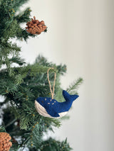 A handcrafted needle-felted humpback whale felt ornament is displayed hanging from a lush green Christmas tree branch adorned with brown pine cones. The gray wool felt ornament features a lighter underside, small white stitch details on its top, and a jute string for easy hanging.