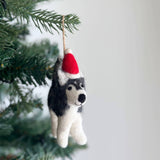 A handcrafted felt husky ornament with a red Christmas hat and white trim hangs by a jute string on a festive Christmas tree. This needle felted husky is crafted from shades of grey and white felt, showcasing its fluffy texture and distinctive markings.