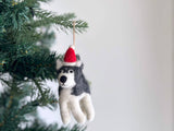 A charming handmade felt husky ornament, adorned with a bright red Christmas hat, hangs on a green Christmas tree branch against a plain white background. The festive husky ornament features a fluffy white belly and paws, contrasted by grey felt on its back and head, with a black nose and simple stitched eyes.