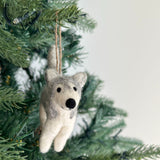 A handcrafted husky felt ornament hangs from a Christmas tree branch, creating a festive atmosphere. This wool felt husky ornament is white with gray markings on its back, ears, and around its small black eyes and nose. A jute string allows it to hang gracefully.