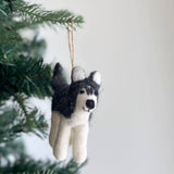 A handcrafted wool felt husky ornament is playfully hung on a Christmas tree branch, set against a simple white background. This black and white husky felt ornament captures distinct markings, featuring upright ears and a smiling face. A jute string connects it to the branch, highlighting its soft, inviting texture and cheerful appearance.