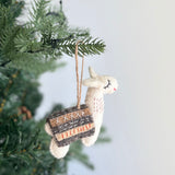 A charming handcrafted wool felt llama ornament hangs from a green Christmas tree branch against a soft white background. This white llama felt ornament features fluffy ears, a red stitched neck, closed eyes, and a small smile. A decorative blanket in dark brown, tan, and white with stitched accents is draped over its back, with a jute string attached for hanging.