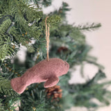 A dusty pink needle felted manatee ornament is displayed in a close-up shot, hanging from a Christmas tree branch. Crafted from soft wool felt, this charming felt manatee ornament features a rounded body, small flippers, and tiny black eyes, suspended by a rustic jute string. The tree branch is decorated with subtle white lights and a natural pine cone, adding to the cozy, festive holiday ambiance.