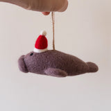 A handcrafted felt manatee ornament is delicately held by a female hand against a light backdrop, evoking a whimsical and cheerful feel. This unique wool felt manatee features a charming red and white Santa hat and includes a jute string for easy hanging.